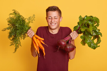 Happy smiling male holding fresh carrot and beets, guy looks satisfied, keeps eyes closed, standing...