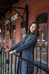 young thoughtful pretty girl in gray coat and turtleneck poses in front of red building. Street style portrait photo session of attractive female, urban photoshoot of beautiful elegant model