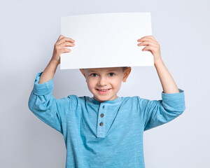 A beautiful boy is holding a clean white sheet of paper on an isolated background, holding it above his head and smiling. © Вера Сидорова