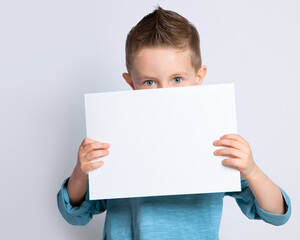 A beautiful boy is holding a clean white sheet of paper on an isolated background, covering the lower half of his face with it. A sly look in a child's blue eyes.