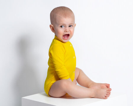 The baby is crying. A baby in a yellow bodysuit sits sideways, screams merrily, winces and makes a face. A child's cry, crying. Portrait of a beautiful baby girl, bright photo, children's emotions. 