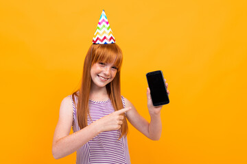 red-haired girl with a festive cap on her head holds a phone with a mockup on a yellow background