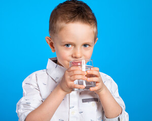 A beautiful European boy drinks water from a glass. Portrait of a child on a blue background. Water in a glass. Thirst, water use, advertising of drinking water.