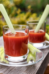 Two glasses of tomato juice with parsley and celery decorations, stand on wooden boards in the garden, morning sunlight shines, shallow depth of field, selective focus. The concept of natural drinks.