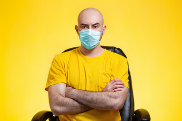 A bald man with glasses and a medical mask is sitting in a chair, his arms crossed in displeasure.Yellow background. Copy space. The concept of self-isolation, quarantine and virus protection