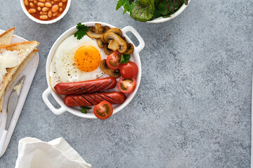 Traditional full English breakfast with fried eggs, sausages, beans, mushrooms, grilled tomatoes on grey background.