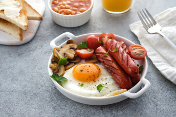 Traditional full English breakfast with fried eggs, sausages, beans, mushrooms, grilled tomatoes on grey background.