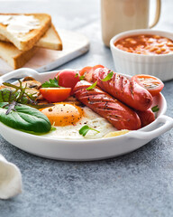 Traditional full English breakfast with fried eggs, sausages, beans, mushrooms, grilled tomatoes on grey background.