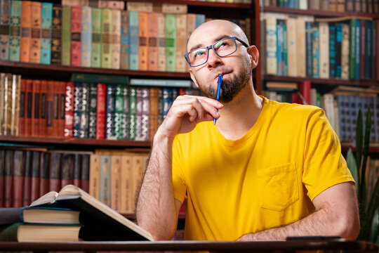 A bald, bearded man with glasses raised a pen to his mouth thoughtfully as he sat in front of his books. There are bookshelves in the background. Concept of distance learning and education