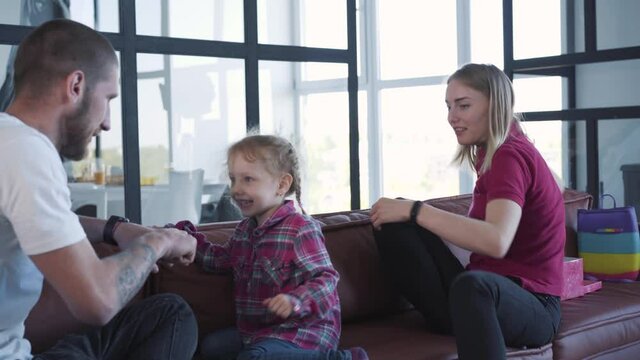 Cute blond girl climbing on sofa and playing with father guess fist as mother trying to comb her pigtails. Young Caucasian family enjoying weekend morning indoors. Leisure, togetherness, lifestyle.