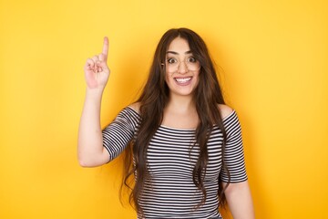 Close-up portrait of charming young woman in casual clothes pointing with one finger up, looking up and smiling, I have an idea, I know the answer or the solution.