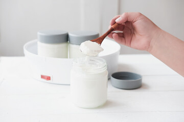 baby's hand holding a spoon with homemade organic yogurt. jars in yogurt maker. automatic yogurt machine to make fermented milk product at home. yogurt or kefir making during quarantine concept. 