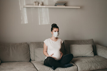 Young beautiful pregnant woman in medical surgical mask and white t-shirt sits on gray couch on...