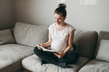 Happy young beautiful pregnant woman sitting on gray couch and reading a book on quarantine at home. Relax, keep calm, rest, coronavirus, virus, isolation, stay at home.