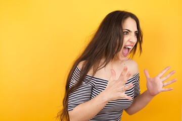 Portrait of young woman with shocked facial expression holding hands near face, screaming and looking sideways at something amazing. Stands against yellow studio Wall.