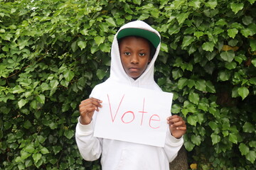 African American Child wearing hoodie and hat holding White paper sign with  word Vote written in red letters bushes background