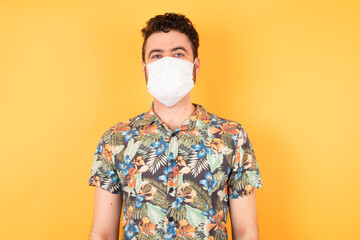 Young caucasian man wearing hawaiian shirt and medical mask  standing over isolated yellow background with a happy and cool smile on face. Lucky person.