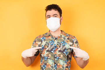 Horizontal shot of cheerful stylish man points at his casual hawaiian shirt and medical mask and safety gloves, stands indoors against concrete wall, being in good mood after going shopping a