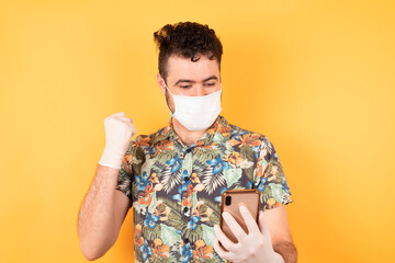 Portrait of caucasian young businessman wearing hawaiian shirt and medical mask and safety gloves holding in hands cell reading browsing isolated on bright vivid shine yellow background