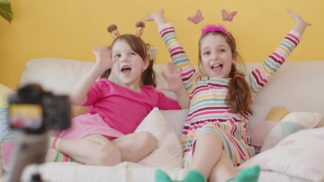 Shooting a Video Blog for Children with Camera on Tripod. Production of Kid's Content. Two Cute Cheerful Little Caucasian Girl Talking and Laughing, Sitting on a Couch. Cozy Interior with Yellow Wall.