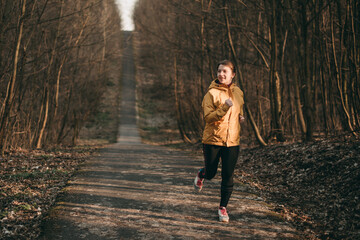 Portrait of young white european blond woman in yellow sports jacket, black leggings and sneakers running in park in spring. Forest, nature, healthy lifestyle, park, sport, sportswear, happy.