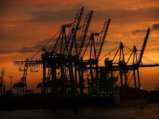 Cargo cranes silhouettes against a dramatic orange sunset sky, Hamburg harbor, Germany.