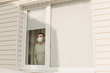 Young brunette white european man at home on quarantine with medical mask stands by window and looks out. Coronavirus, illness, infection, quarantine, surgical bandage. photo from the window street