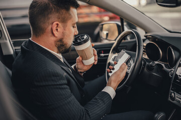 Handsome caucasian businessman driver inside in vehicle  with coffee and gadget and check mail and talking with partners