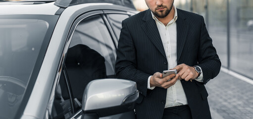 Handsome businessman with smartphone near vehicle on office parking