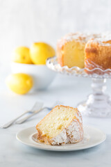 Front view of a slice of lemon bundt cake with the cake on a cake stand, forks and a bowl of lemons in soft focus.