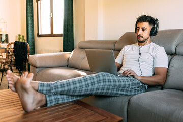 A man sitting in house clothes on the couch working remotely. The young man has the computer on his legs and the headphones on his head. Working remotely from home. The boy plays video games online.