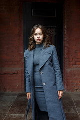 Fototapeta premium young thoughtful pretty girl in gray coat and turtleneck poses in front of red building. Street style portrait photo session of attractive female, urban photoshoot of beautiful elegant model