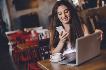 Business woman using phone, working on a laptop and drinking coffee in a cafe.