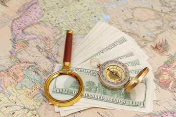 A bundle of American dollars lies with a compass and a magnifying glass on the world map