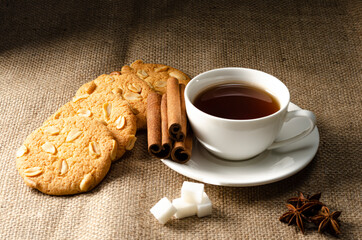 Cookies with peanuts and tea in a white cup on a saucer with sugar cubes, anise stars and a cinnamon stick. On the background of burlap