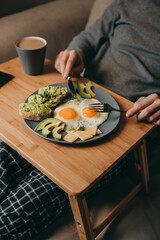 The guy sits on the couch with breakfast table and eats breakfast on a plate of fried eggs, bread, avocado toast, cheese and coffee, a smartphone on the table.