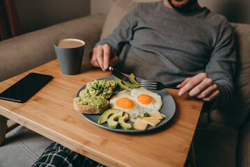 The guy sits on the couch with breakfast table and eats breakfast on a plate of fried eggs, bread, avocado toast, cheese and coffee, a smartphone on the table.