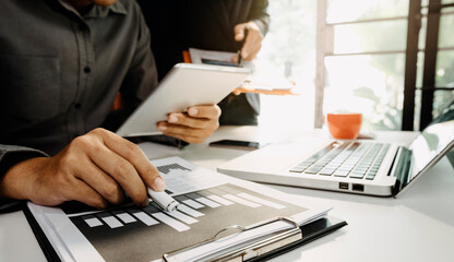 Businessman hand using laptop and tablet with social network diagram and two colleagues discussing data on desk as concept in morning light.