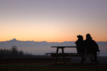 Obraz na płótnie Canvas silhouettes of a Couple sitting in a bench enjoying the wonderful color of the sky at dusk, in the background the top of the pink mountain, Italy.