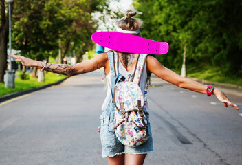 Outdoors lifestyle portrait of trendy pretty girl going ahead with pink pennyboard behind her back. Walking, enjoying sun and listening music in hheadphones. Wearing stylish sunglasses, denim overalls