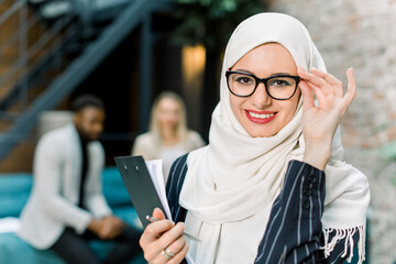 Charming arabian Muslim business woman in white hijab holding folder of papers and posing on camera with happy smile, standing in modern loft office on the background of multiethnic colleagues