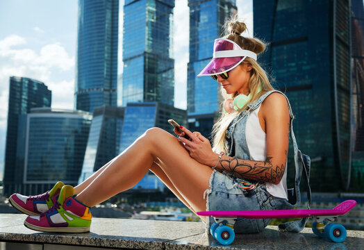Summer lifestyle image of trendy pretty girl sitting on quay on the big city background. Smiling, texting a message,  enjoying sun. Wearing stylish sunglasses, denim overalls, headphones, plastic cap