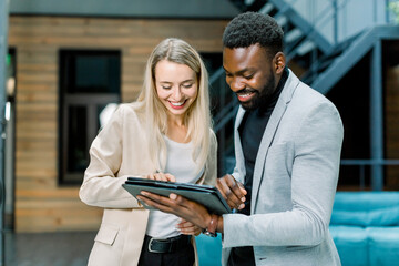 Front view of successful smiling young multiracial business people, Caucasian woman and African bearded man, standing in the office room and talking each other, looking at tablet pc