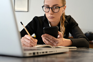 Young woman with eyeglasses, headphones, laptop studying working online writing in note pad....