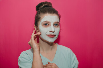 Young beautiful girl with big blue eyes smiling hands near face, with a white clay mask on her face...