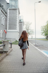 Rear view woman with leather backpack wearing down jacket goes on city street at morning or...