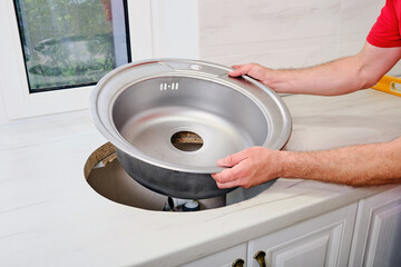 mans hands installs new round single stainless steel sink into a cut-out hole in the kitchen worktop. 