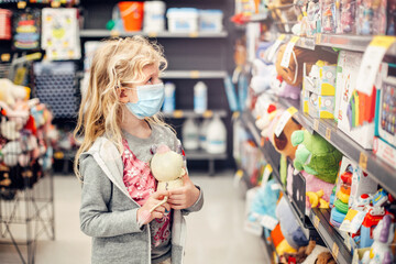A new normal. Caucasian blonde girl in sanitary face mask shopping at toy store. Child wearing...