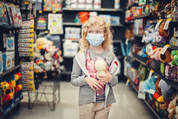 Fototapeta na wymiar A new normal. Caucasian blonde girl in sanitary face mask shopping at toy store. Child wearing protective mask against coronavirus. Safety, health protection during covid-19 quarantine.