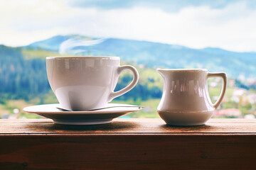 Plans after quarantine. Coffee with milk in ceramic cup with small milk jug on old wooden window sill with summer mountains background.